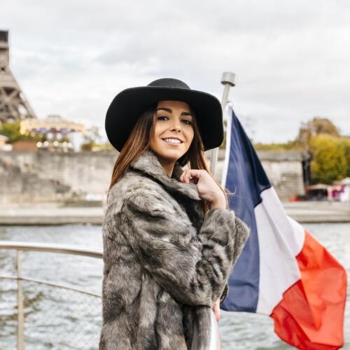 Jeune femme posant près d’un drapeau français avec la tour Eiffel floue en arrière-plan.