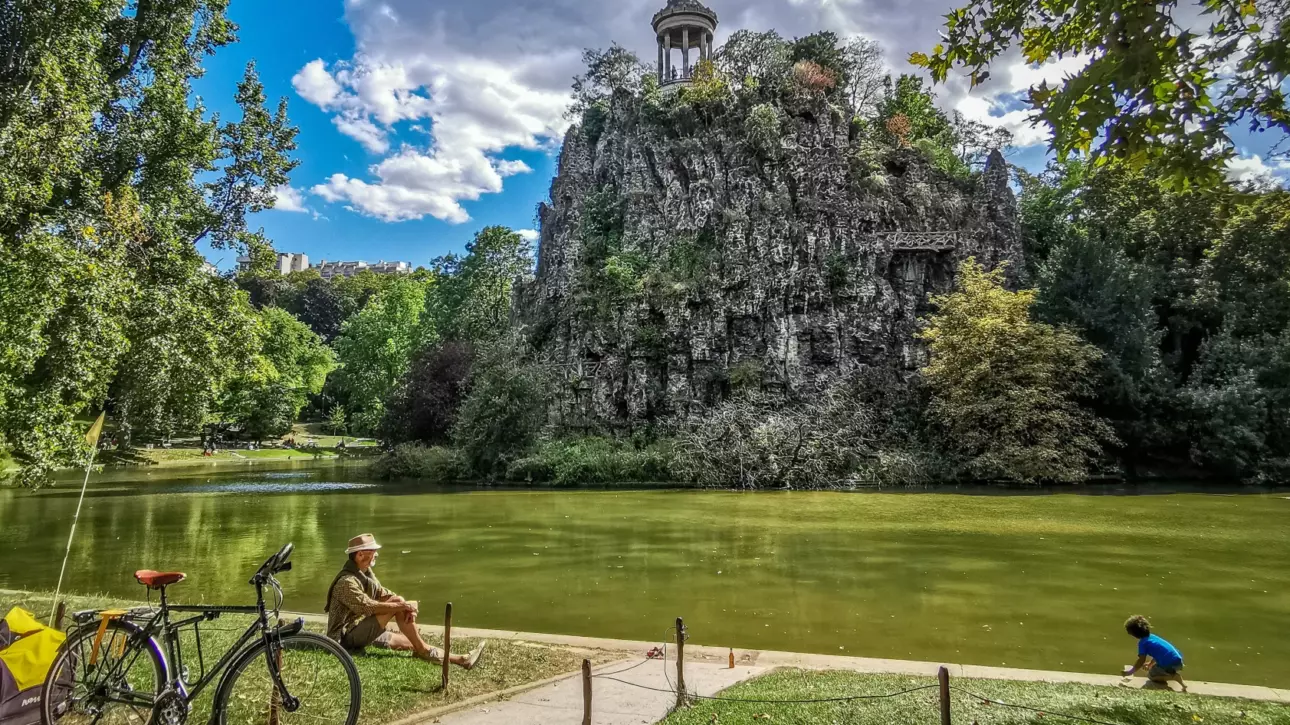 Un homme assis au parc des Buttes-Chaumont avec un enfant à sa droite, près d'une rivière et d'une colline.